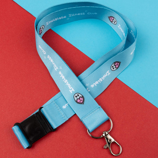 Light blue lanyard with black safety buckle and metal clasp, featuring the 'Invisible Illness Club' text and a charming ghost logo. Designed to raise awareness for invisible illnesses, it is shown against a red and blue geometric background.