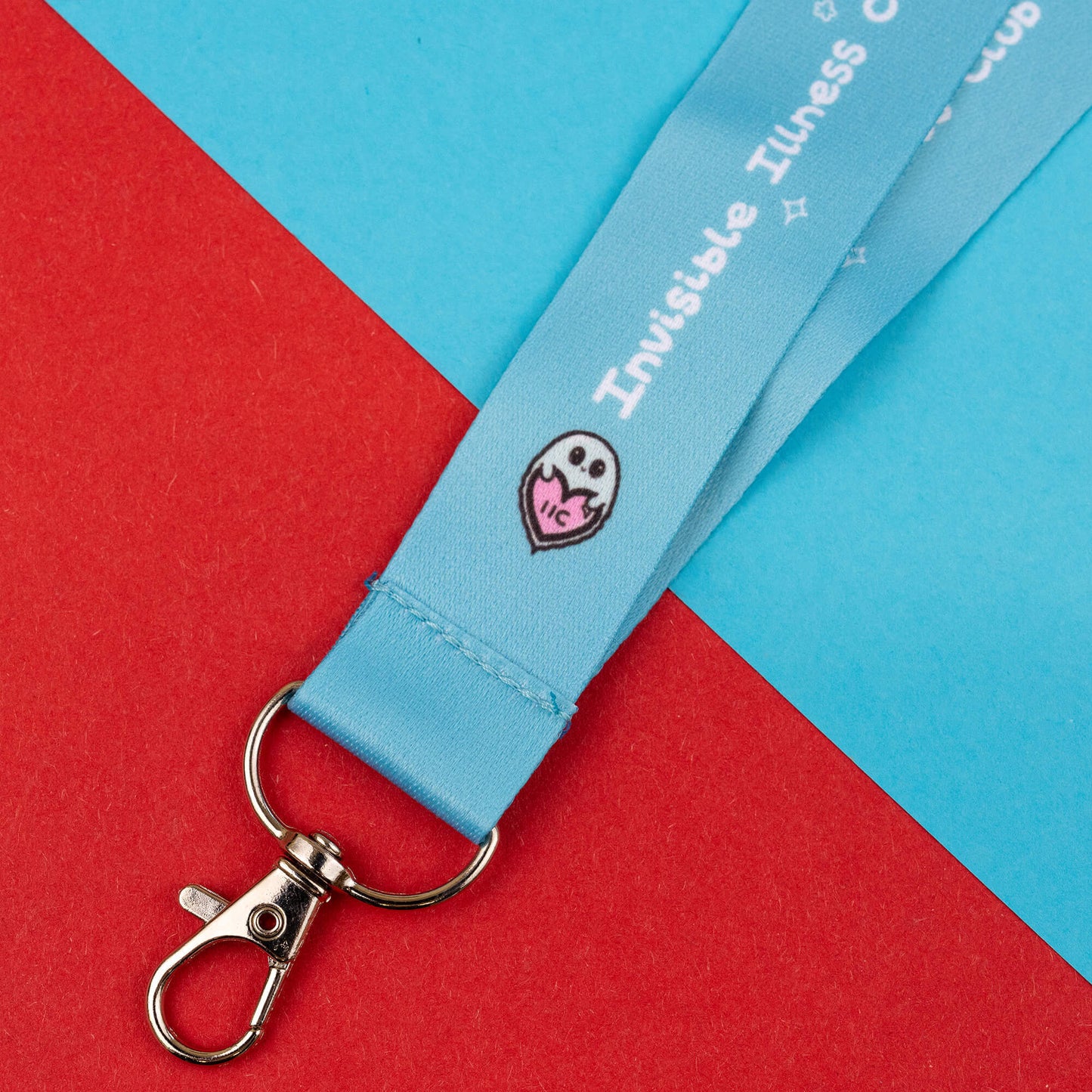 Close-up of a lanyard designed to raise awareness for invisible illnesses. The light blue lanyard features the text 'Invisible Illness Club' and a cute logo of a ghost with a heart. It has a metal clasp and is laid out on a red and blue geometric background.