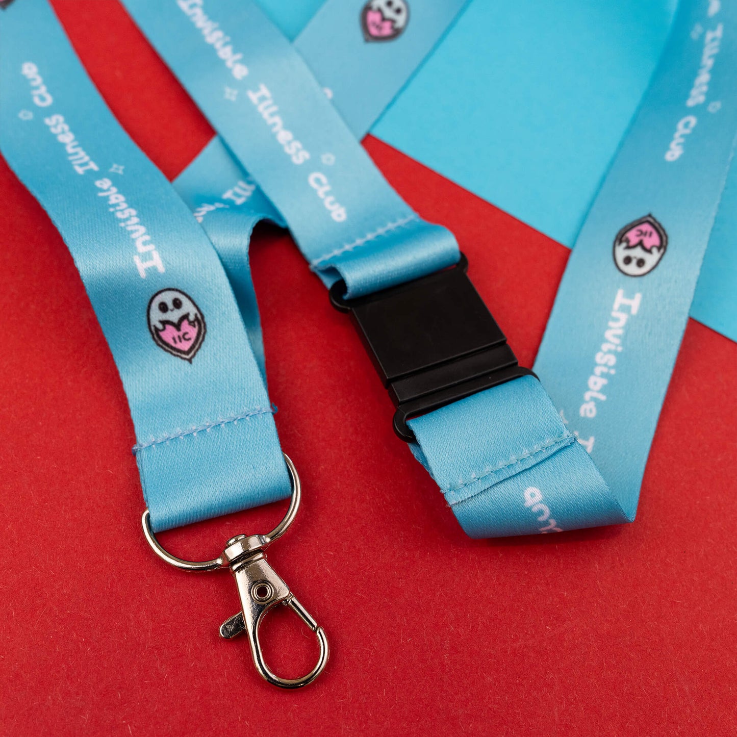 Lanyard with a black buckle and metal clasp, displaying the 'Invisible Illness Club' text and a cute ghost logo. This light blue lanyard is designed to raise awareness for invisible illnesses and is arranged on a red and blue geometric background.