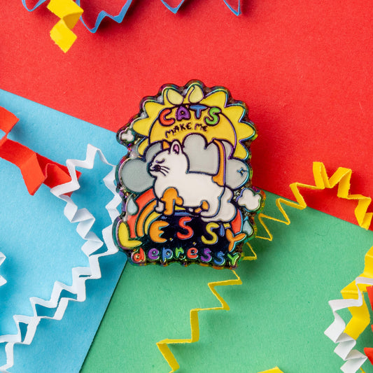 A close-up shot of an Innabox enamel pin featuring a white cat walking under a sun and rainbow, with the text “Cats make me lessy depressy.” The pin sits on a colourful background of red, green, and blue with curly paper decorations around it. This disability-inspired accessory is designed to bring a sense of positivity to those with chronic illnesses or hidden disabilities.