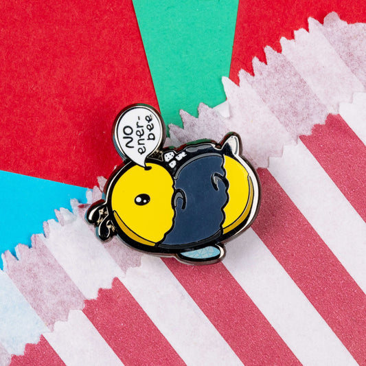 Low Energy Bee Enamel Pin resting on a red and white striped paper bag on a blue, red and teal background. The enamel pin is a cute yellow and black bee lying on it's wings with its arms, legs, little belly and stinger in the air. The bee has a speech bubble coming from it's mouth with 'NO ener-bee' written inside in black writing. Hand drawn design made to raise awareness for chronic fatigue.