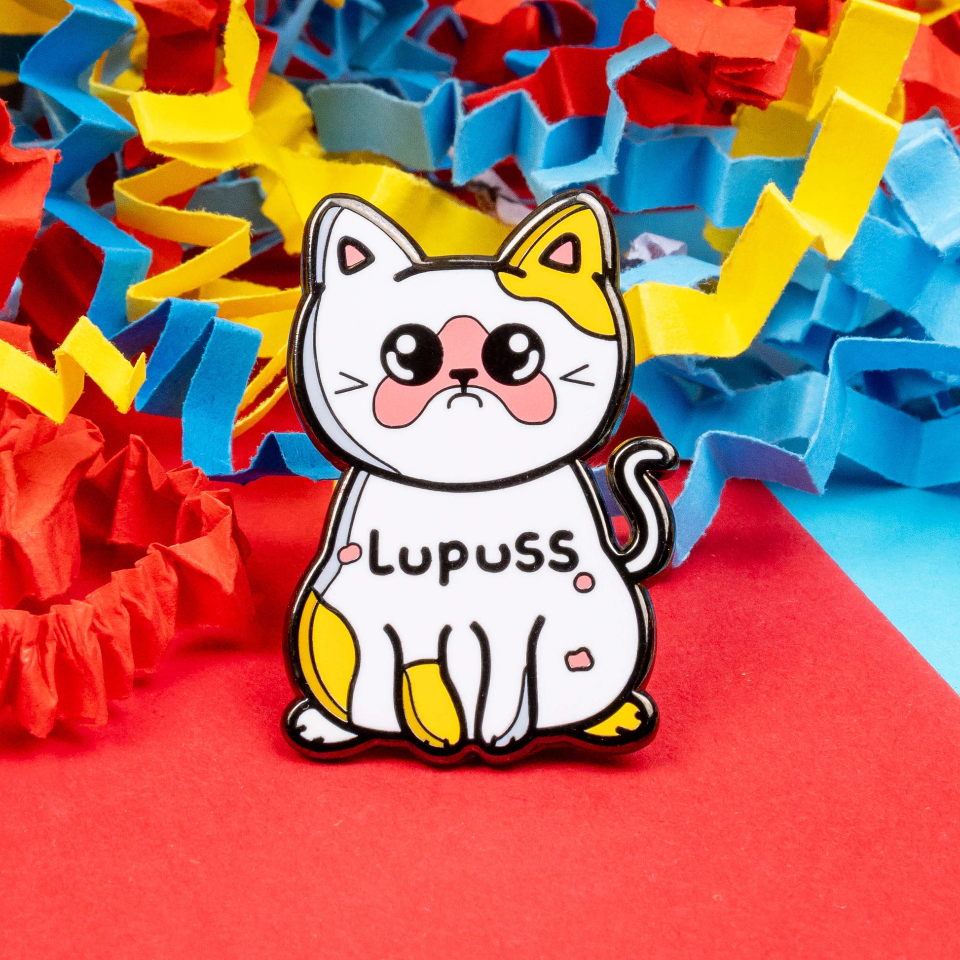 Lupuss Cat Enamel Pin - Lupus on a red, blue and yellow card confetti background. The cat shape enamel pin is orange and white with a sad expression, pink patches all over and black text across its middle reading 'lupuss'. The hand drawn design is raising awareness for lupus.