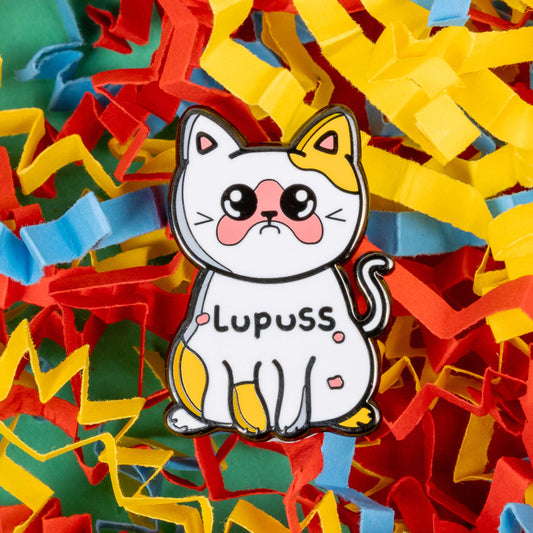 Lupuss Cat Enamel Pin - Lupus on a red, blue and yellow card confetti background. The cat shape enamel pin is orange and white with a sad expression, pink patches all over and black text across its middle reading 'lupuss'. The hand drawn design is raising awareness for lupus.