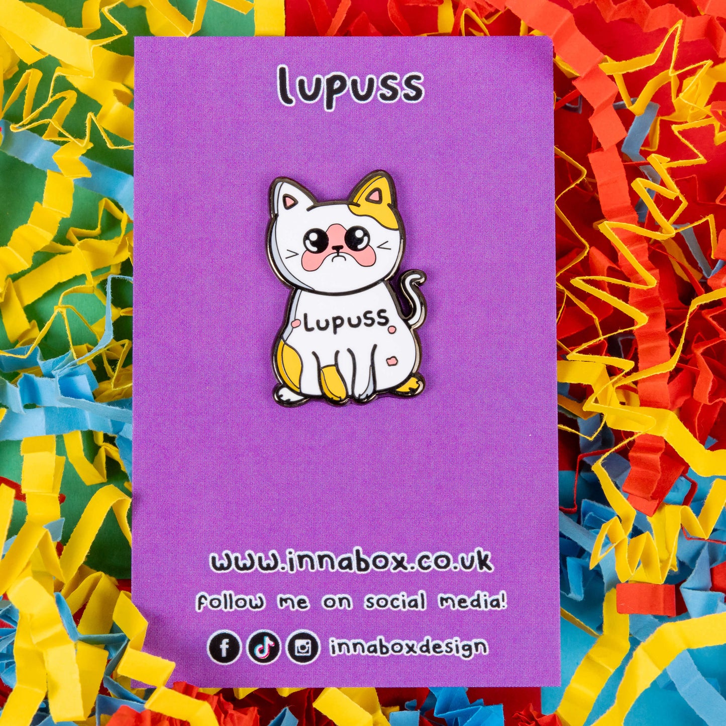 Lupuss Cat Enamel Pin - Lupus on it's purple backing card on a red, blue and yellow card confetti background. The cat shape enamel pin is orange and white with a sad expression, pink patches all over and black text across its middle reading 'lupuss'. The hand drawn design is raising awareness for lupus.