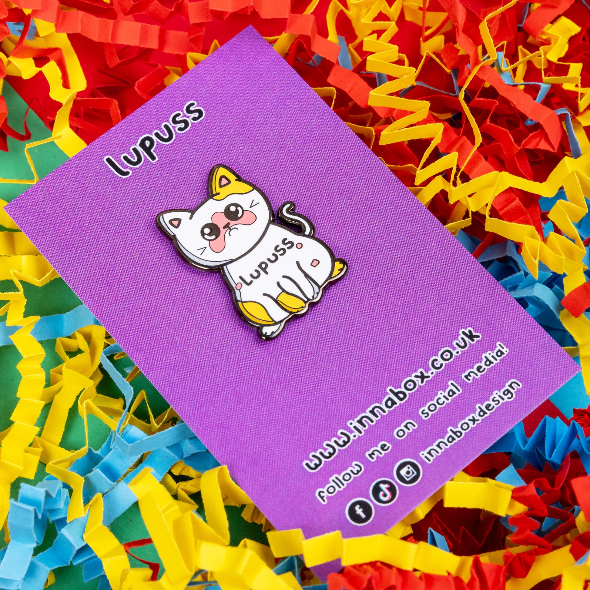 Lupuss Cat Enamel Pin - Lupus on it's purple backing card on a red, blue and yellow card confetti background. The cat shape enamel pin is orange and white with a sad expression, pink patches all over and black text across its middle reading 'lupuss'. The hand drawn design is raising awareness for lupus.