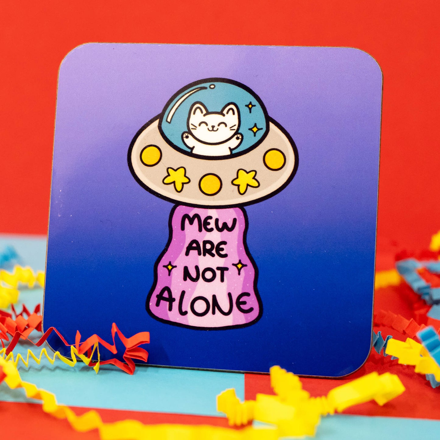 Close-up of the Cat UFO Mew Are Not Alone Coaster by Innabox. The square coaster has rounded corners and a gradient blue background. The design features a cute white cat sitting inside a grey UFO with yellow lights and star details. A pink sparkly beam shines down with the words "Mew Are Not Alone" written in bold, playful letters. Surrounding the coaster are colourful red, blue, and yellow crinkle paper decorations.