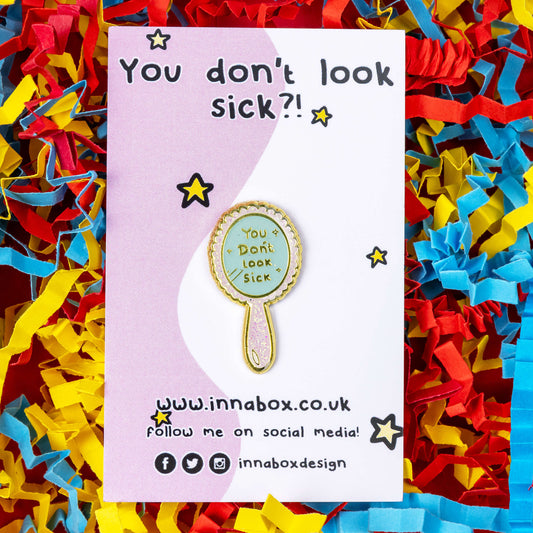 You Don't Look Sick Mirror Enamel Pin on it's pink and white backing card with gold stars on a red, yellow and blue coloured card confetti background. The hand mirror shaped baby pink glittery enamel pin has gold text on it that reads 'you don't look sick'. The hand drawn design is made to raise awareness for invisible illnesses