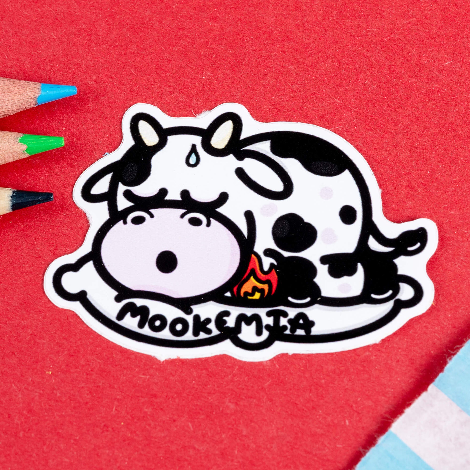 Close-up of the Mookemia Cow Sticker by Innabox. The sticker features a tired, spotted black-and-white cow lying down with a sweat drop on its forehead and flushed pink cheeks. A small flame icon is near its side, symbolising the fatigue often associated with leukemia. The word "Mookemia" is written in bold, playful black letters along the bottom. The background is red, with coloured pencils nearby.