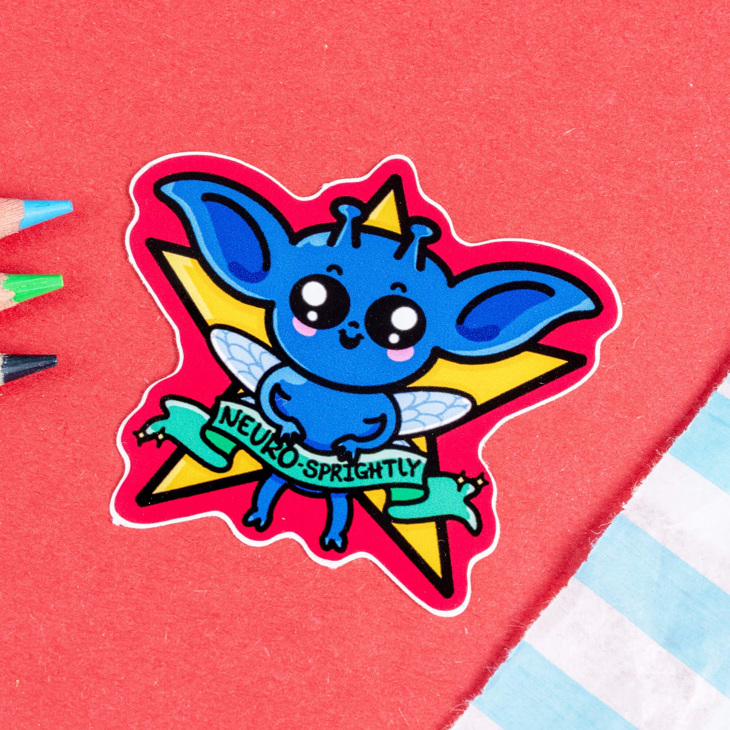 Close-up of the Neuro-Sprightly Sprite Sticker by Innabox. The sticker features a vibrant blue sprite with large, pointy ears, tiny antennae, and glittery fairy wings. It has big, sparkling eyes, rosy cheeks, and a playful smile. Behind the sprite is a yellow star with a pink outline, and a green ribbon banner reading "Neuro-Sprightly." Set against a red background with coloured pencils nearby.