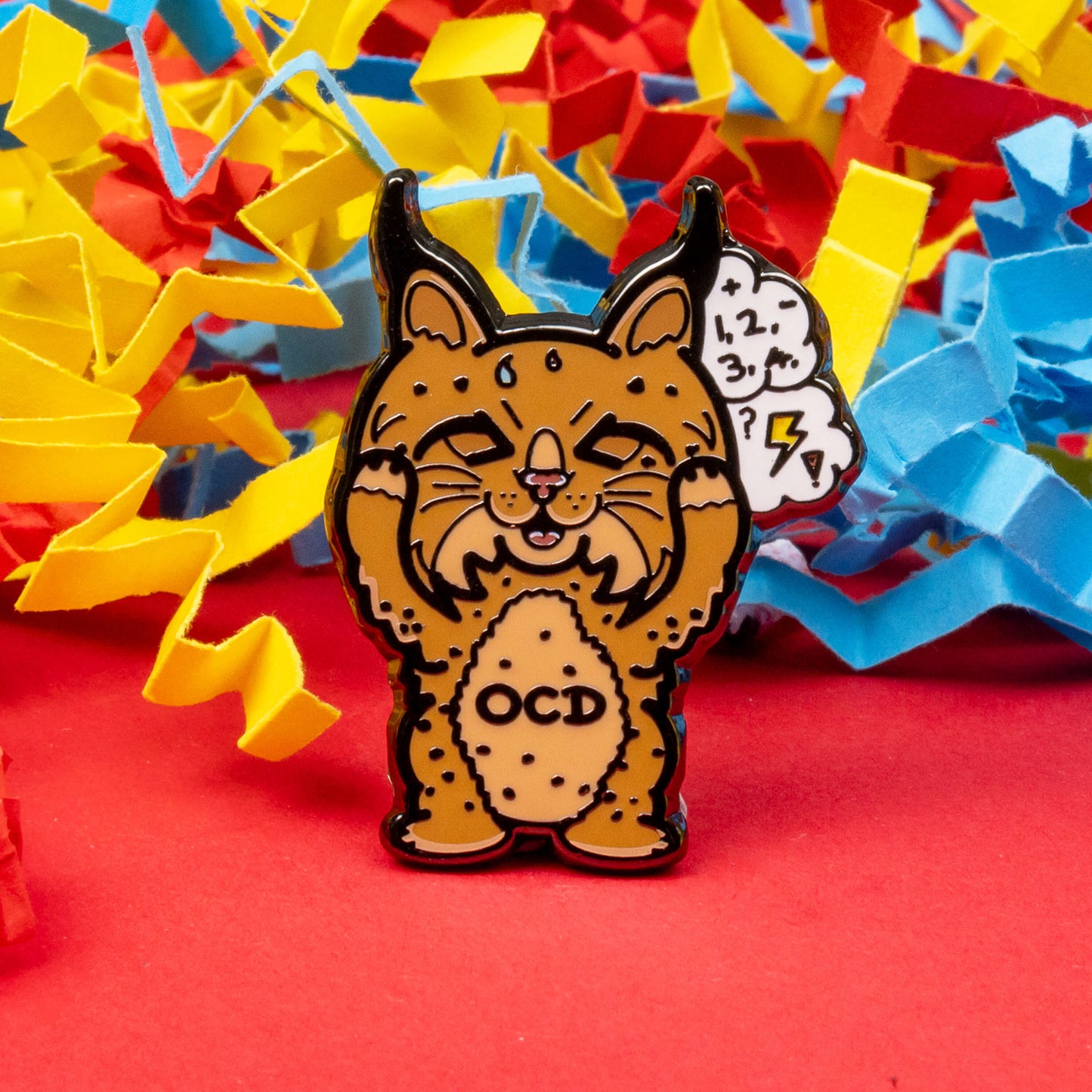 Bobsessive Compulsive Disorder Enamel Pin - OCD on a red, yellow and blue coloured card confetti background. The bobcat shape pin is brown with a stressed expression clutching its face with a thought bubble of numbers, question marks and lightning bolts. Across its tummy reads OCD. The pin was designed to raise awareness of obsessive compulsive disorder.