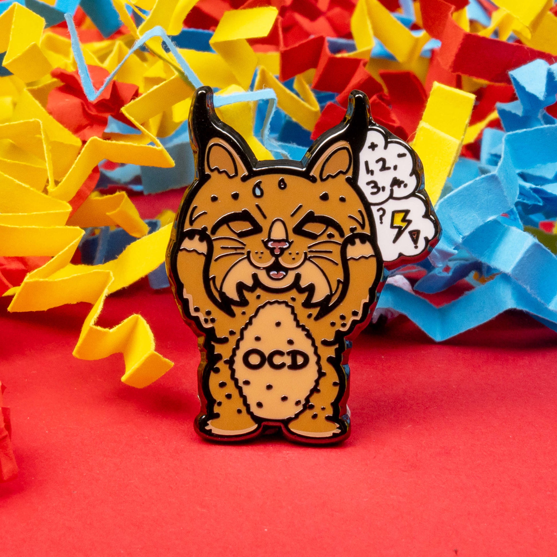 Bobsessive Compulsive Disorder Enamel Pin - OCD on a red, yellow and blue coloured card confetti background. The bobcat shape pin is brown with a stressed expression clutching its face with a thought bubble of numbers, question marks and lightning bolts. Across its tummy reads OCD. The pin was designed to raise awareness of obsessive compulsive disorder.