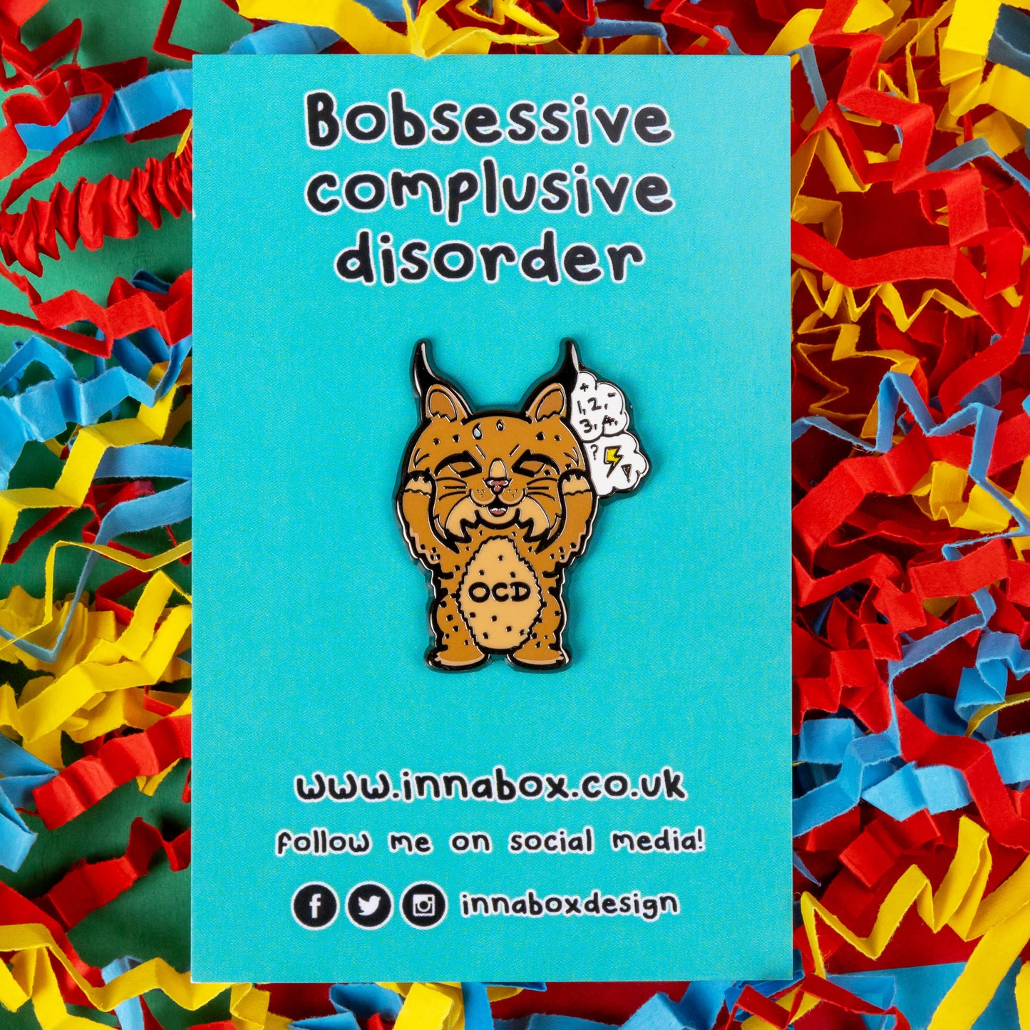 Bobsessive Compulsive Disorder Enamel Pin - OCD on blue backing card with innabox social media info on a red background with coloured card confetti. The bobcat shape pin is brown with a stressed expression clutching its face with a thought bubble of numbers, question marks and lightning bolts. Across its tummy reads OCD. The pin was designed to raise awareness of obsessive compulsive disorder.