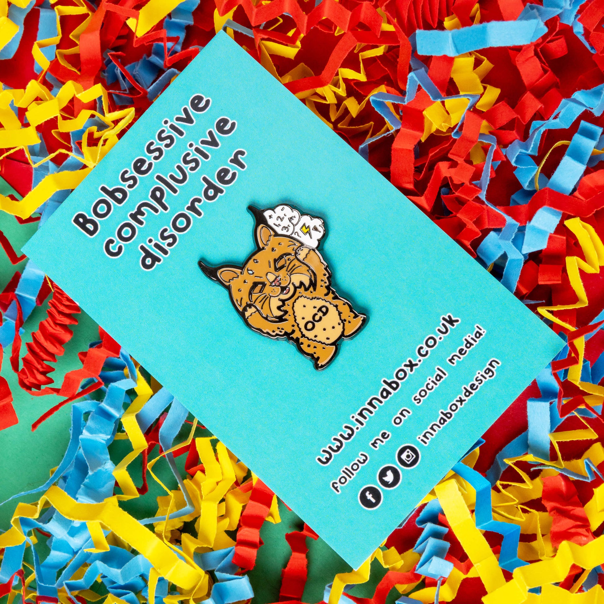 Bobsessive Compulsive Disorder Enamel Pin - OCD on blue backing card with innabox social media info on a red background with coloured card confetti. The bobcat shape pin is brown with a stressed expression clutching its face with a thought bubble of numbers, question marks and lightning bolts. Across its tummy reads OCD. The pin was designed to raise awareness of obsessive compulsive disorder.