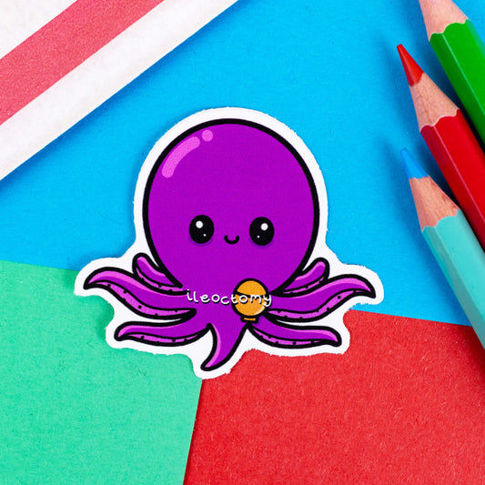 The Ileoctomy Octopus Sticker - Ileostomy on a red, blue and green background with colouring pencils and red stripe candy bag. The purple cute octopus sticker is smiling with a stoma bag and white text across its body reading 'ileoctomy'. The hand drawn design is raising awareness for Ileostomy.