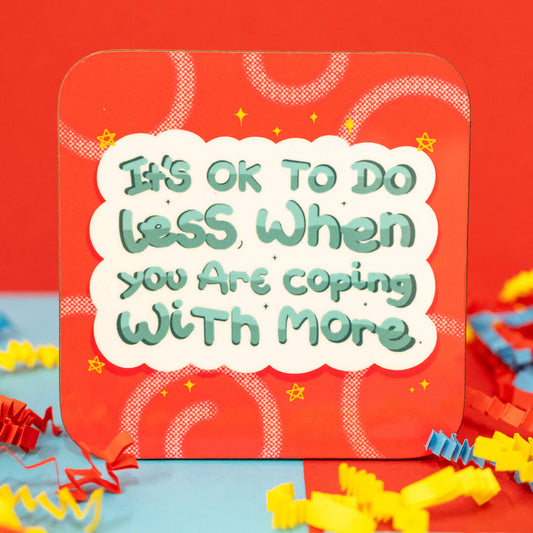 Close-up of the It's Ok To Do Less Coaster by Innabox. The square coaster has rounded corners and a bold red background with swirly patterns, tiny stars, and a small rainbow in the top right. The design features a fluffy white speech bubble with green text reading, "It's OK to do less when you are coping with more." Colourful red, blue, and yellow crepe paper surrounds the coaster.