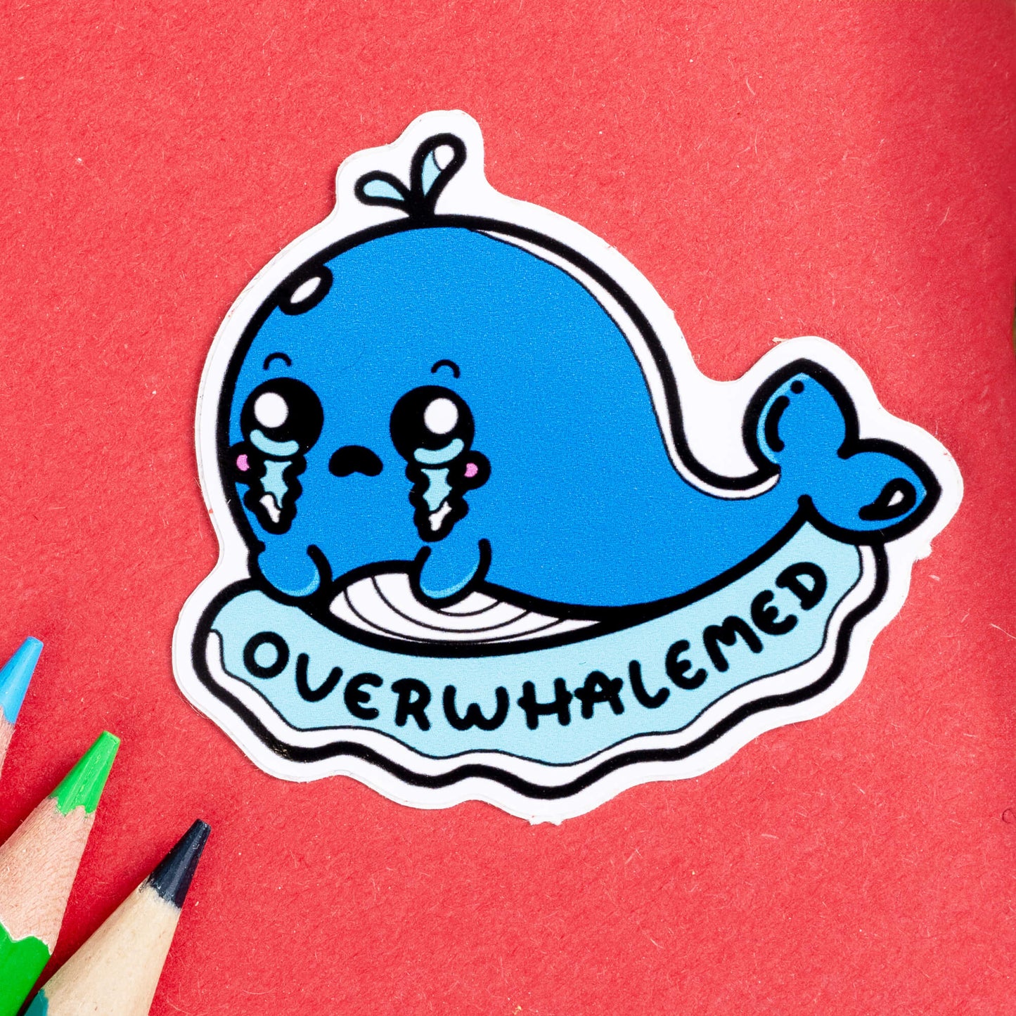 Close-up of the Overwhalemed Whale Sticker by Innabox. The sticker features a teary-eyed blue whale with a tiny water spout on its head. Its cheeks are flushed, and large tears stream down its face as it rests on a light blue wave with the word "Overwhalemed" written across the bottom in bold, playful black letters. The background is red, with coloured pencils nearby.