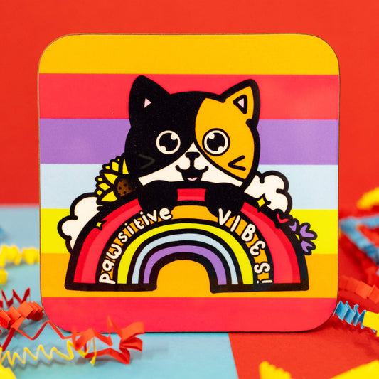 Close-up of the Pawsitive Vibes Cat Coaster by Innabox. The square coaster has rounded corners and a vibrant rainbow-striped background. The design features an adorable calico cat with one black side and one orange side, peeking over a bold red rainbow with the words "Pawsitive Vibes!" written across it. Sunflowers, lavender, and fluffy clouds add extra charm. Colourful crinkle paper surrounds the coaster.