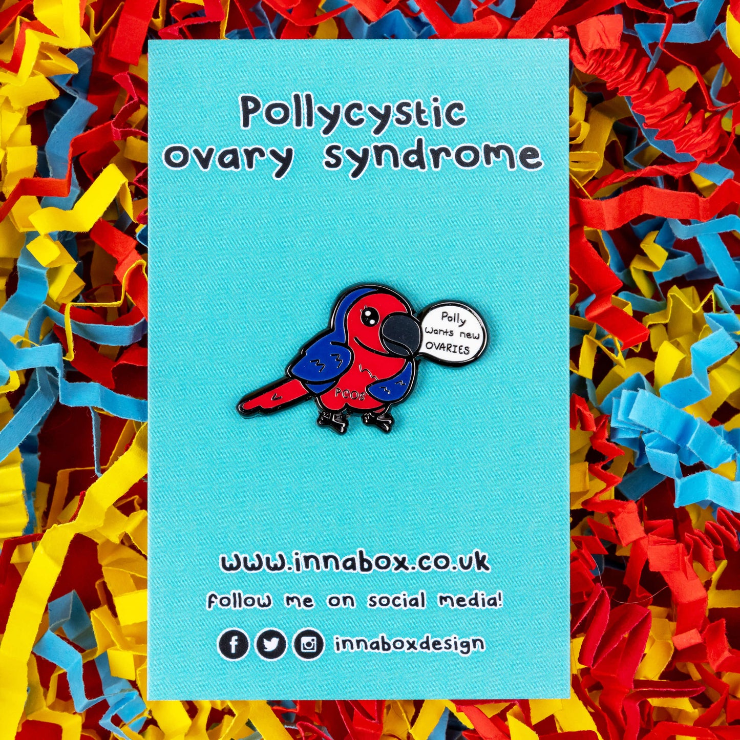 The Pollycystic Ovary Syndrome Parrot Enamel Pin - Polycystic Ovary Syndrome PCOS on blue backing card on a red, blue and yellow card confetti backdrop. The blue and red parrot pin badge is smiling with a speech bubble reading 'polly wants new ovaries', across its middle reads 'PCOS'. The hand drawn design is raising awareness for Polycystic Ovary Syndrome PCOS.