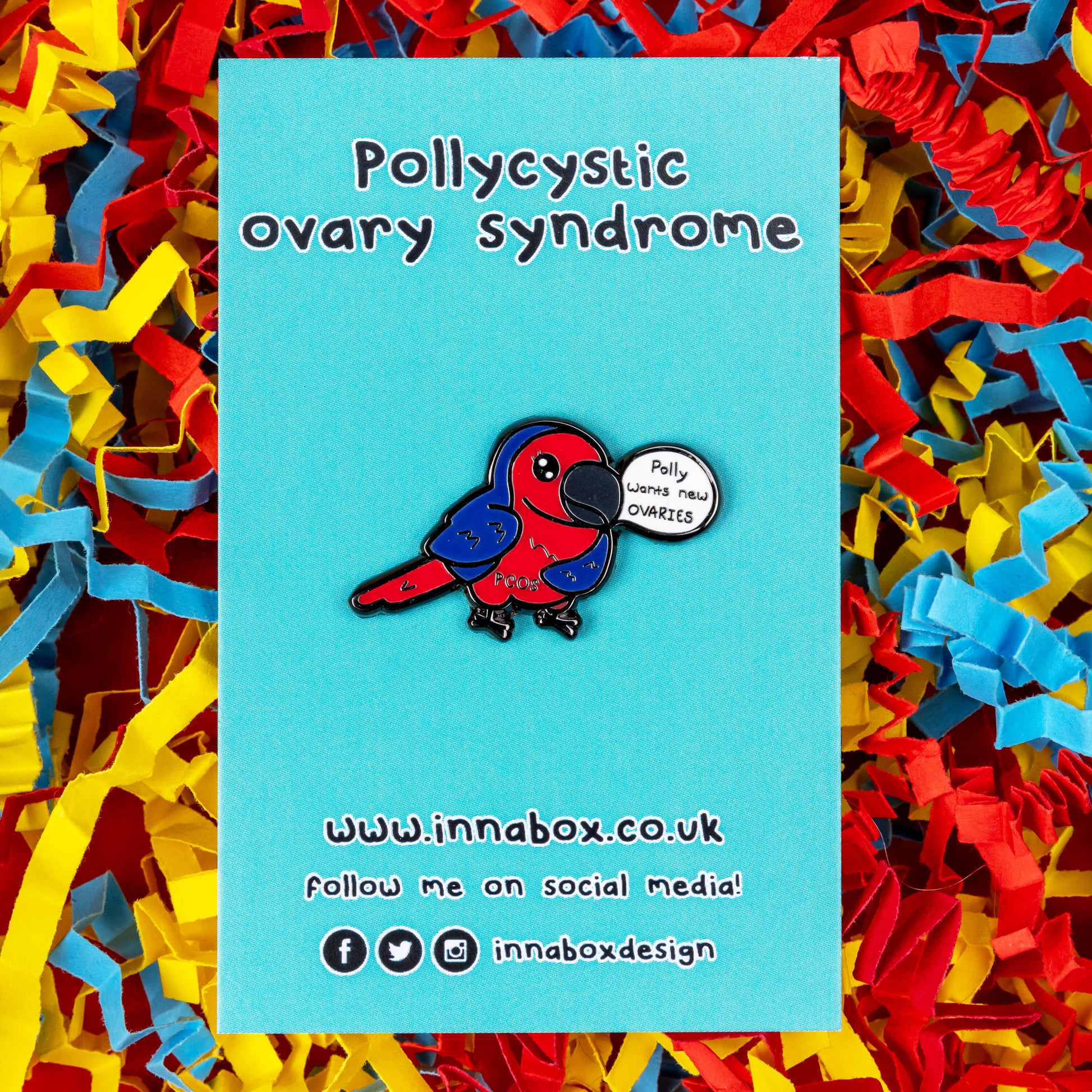 The Pollycystic Ovary Syndrome Parrot Enamel Pin - Polycystic Ovary Syndrome PCOS on blue backing card on a red, blue and yellow card confetti backdrop. The blue and red parrot pin badge is smiling with a speech bubble reading 'polly wants new ovaries', across its middle reads 'PCOS'. The hand drawn design is raising awareness for Polycystic Ovary Syndrome PCOS.