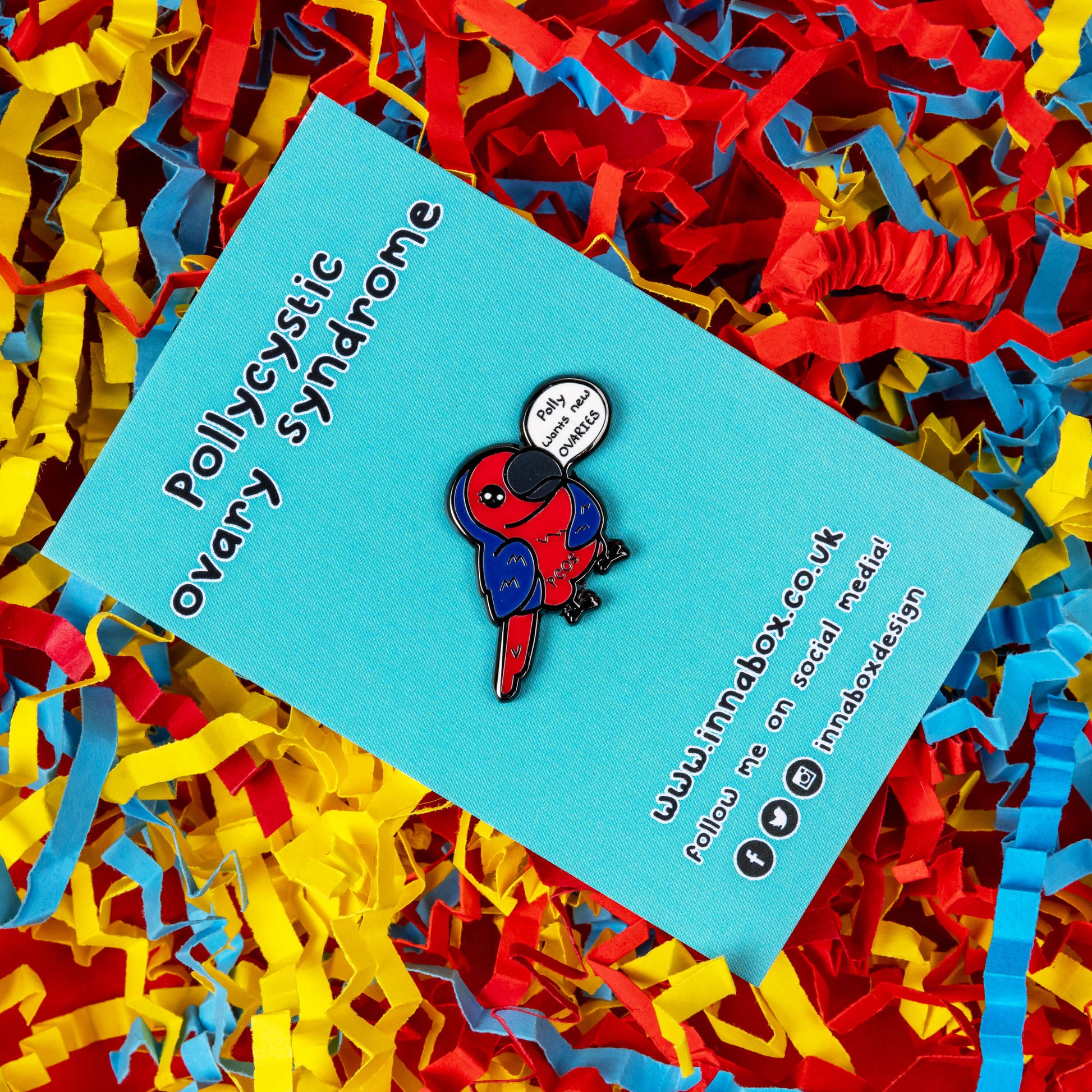 The Pollycystic Ovary Syndrome Parrot Enamel Pin - Polycystic Ovary Syndrome PCOS on blue backing card on a red, blue and yellow card confetti backdrop. The blue and red parrot pin badge is smiling with a speech bubble reading 'polly wants new ovaries', across its middle reads 'PCOS'. The hand drawn design is raising awareness for Polycystic Ovary Syndrome PCOS.