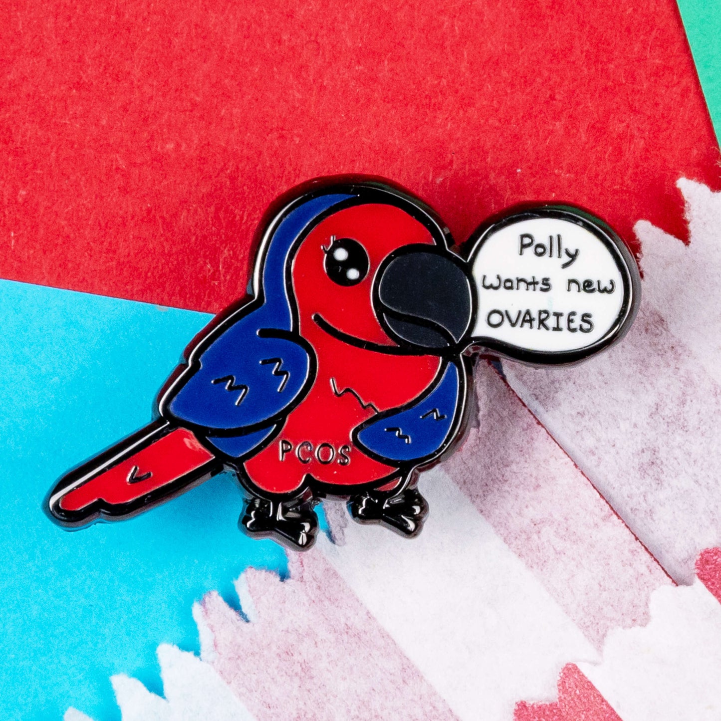 The Pollycystic Ovary Syndrome Parrot Enamel Pin - Polycystic Ovary Syndrome PCOS resting on a red and white striped paper bag on a red and blue background. The blue and red parrot pin badge is smiling with a speech bubble reading 'polly wants new ovaries', across its middle reads 'PCOS'. The hand drawn design is raising awareness for Polycystic Ovary Syndrome PCOS.