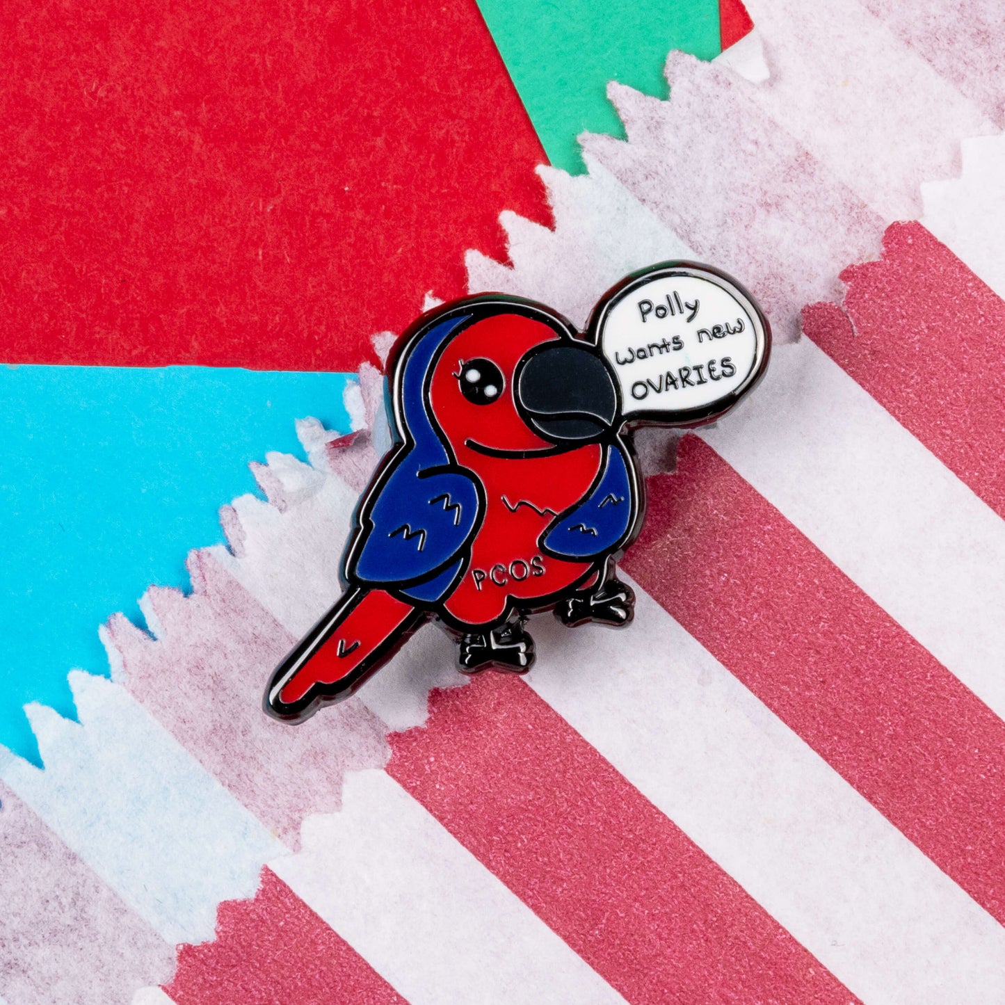 The Pollycystic Ovary Syndrome Parrot Enamel Pin - Polycystic Ovary Syndrome PCOS resting on a red and white striped paper bag on a red and blue background. The blue and red parrot pin badge is smiling with a speech bubble reading 'polly wants new ovaries', across its middle reads 'PCOS'. The hand drawn design is raising awareness for Polycystic Ovary Syndrome PCOS.