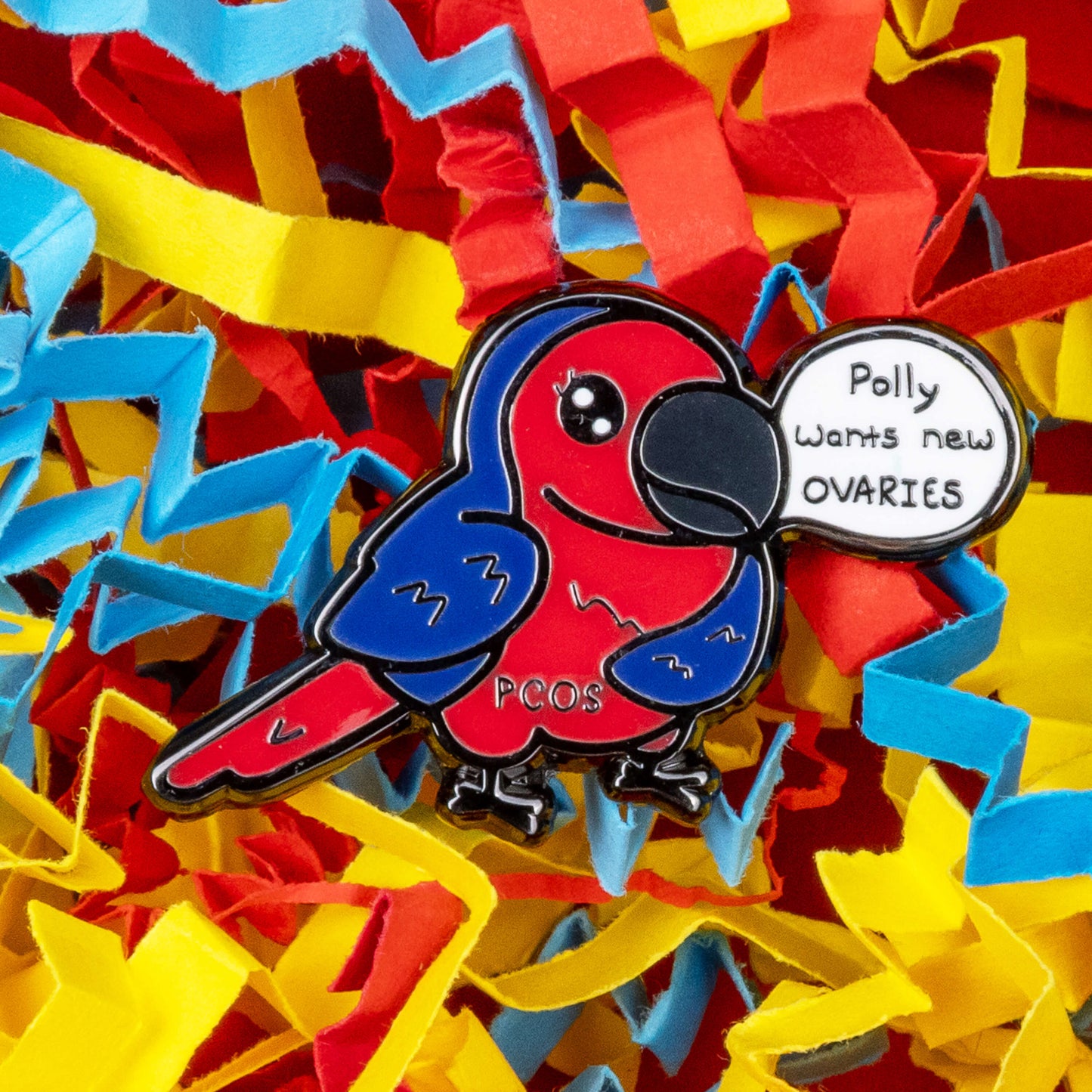 The Pollycystic Ovary Syndrome Parrot Enamel Pin - Polycystic Ovary Syndrome PCOS on a red, blue and yellow card confetti backdrop. The blue and red parrot pin badge is smiling with a speech bubble reading 'polly wants new ovaries', across its middle reads 'PCOS'. The hand drawn design is raising awareness for Polycystic Ovary Syndrome PCOS.