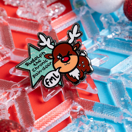 Close-up of Innabox's 'Rudolph the Chronic Pain-deer' pin, showcasing a playful and empowering design on a colourful red and blue background with snowflake decorations, celebrating resilience in chronic illness.