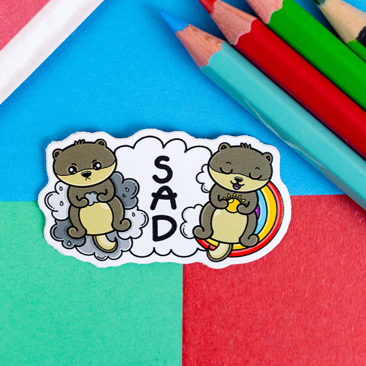 The Seasonal Affective Otter Sticker - Seasonal Affective Disorder SAD on a red, blue and green background with colouring pencils and red stripe candy bag. The cloud shaped sticker has two otters on either side, one sad on a raincloud clutching a raincloud and the other smiling on a rainbow clutching a sunshine. In the middle is the initials 'SAD'. The hand drawn design is raising awareness for Seasonal Affective Disorder.