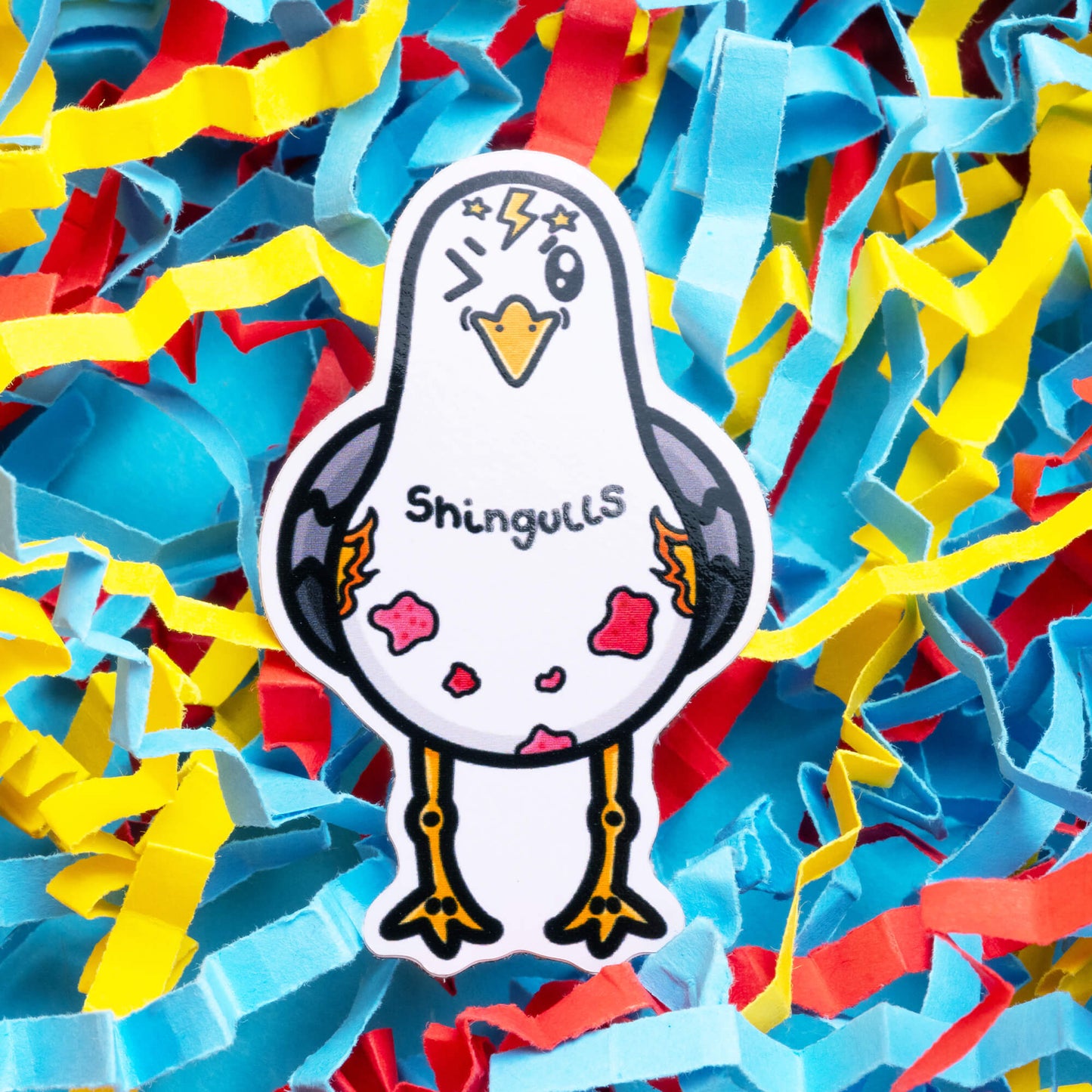 Innabox Shingulls sticker showcasing a cute seagull with pink patches and lightning bolts, symbolising shingles. The 'Shingulls' text is displayed on the seagull, set against a colourful shredded paper background, highlighting this uplifting product for chronic illness awareness.