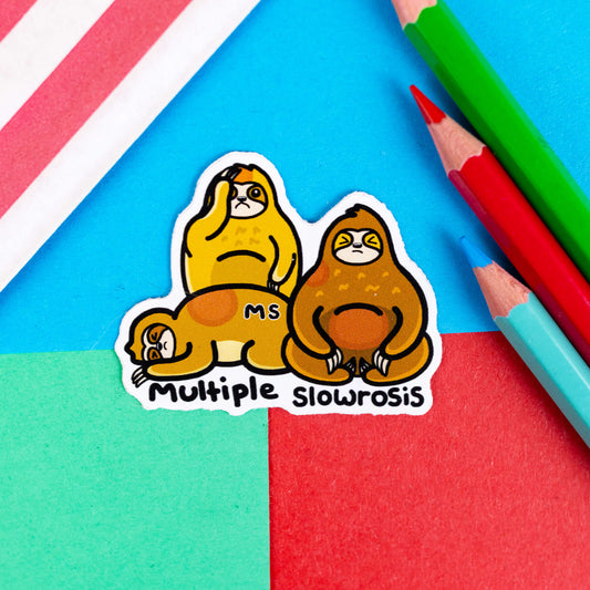 The Multiple Slowrosis Sloth Sticker - Multiple Sclerosis / MS on a red, blue and green background with colouring pencils and red stripe candy bag. A sticker of three sad sloths, two brown and one yellow, with red patches all over their body. Across one in black text reads 'MS' and underneath in black reads 'multiple slowrosis'. The hand drawn design is raising awareness for Multiple Sclerosis / MS.