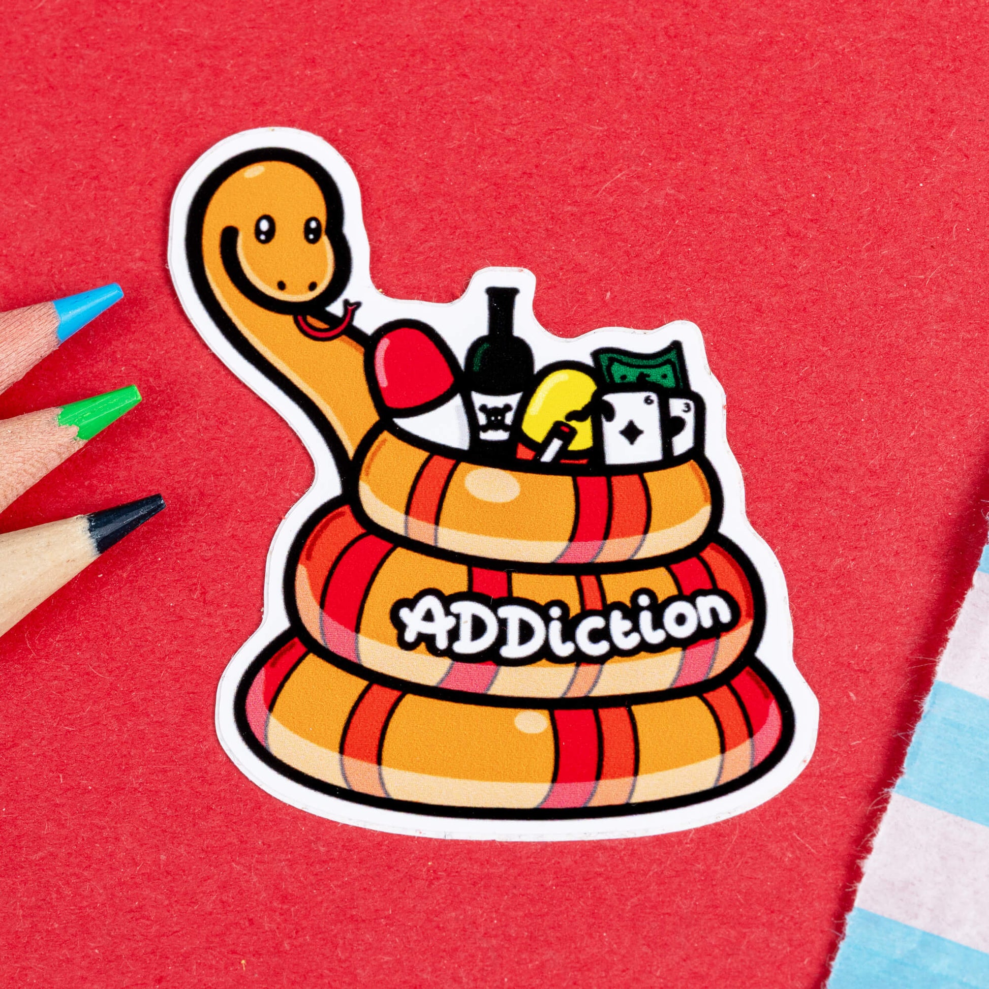 Close-up of the Addiction Adder Snake Sticker by Innabox. The sticker features an orange and red striped snake coiled up with a smiling face. Inside its coils are objects representing addiction struggles, including a wine bottle, playing cards, cash, pills, and a balloon. The word "ADDIction" is written across the bottom in bold, playful white letters. The background is red, with coloured pencils nearby.