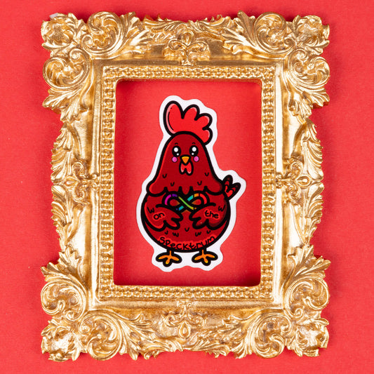 A red cartoon chicken sticker with a rainbow infinity symbol on its belly, holding the symbol with its wings. The text "on the specktrum" is written on the chicken's body. The sticker is framed by an ornate, gold decorative frame and placed on a solid red background. Hand drawn design created to raise awareness for ASD and neurodivergence