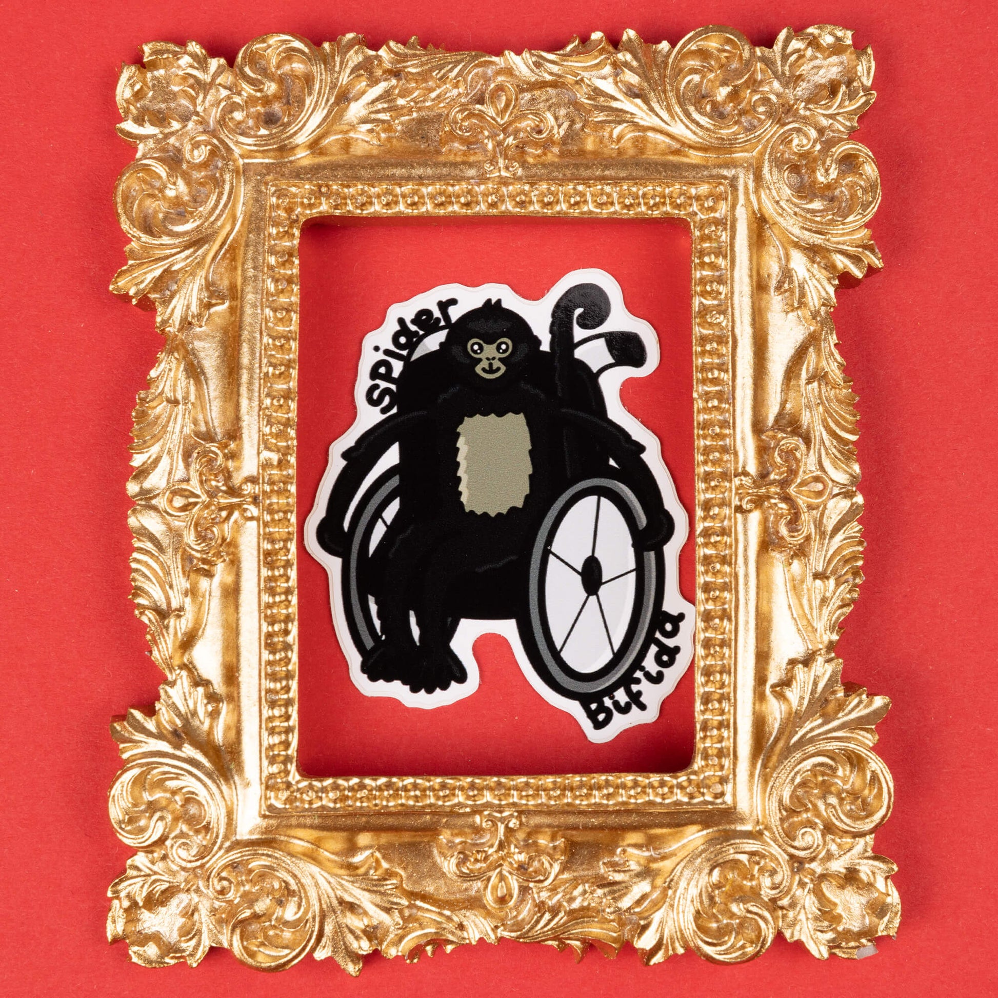 A cartoon sticker of a black spider monkey sitting in a wheelchair. The monkey has a light-colored patch on its chest and a smiling face. The text "Spider Bifida" is written on the sticker. The sticker is framed by an ornate, gold decorative frame and placed on a solid red background. Hand drawn design created created to raise awareness for spina bifida.