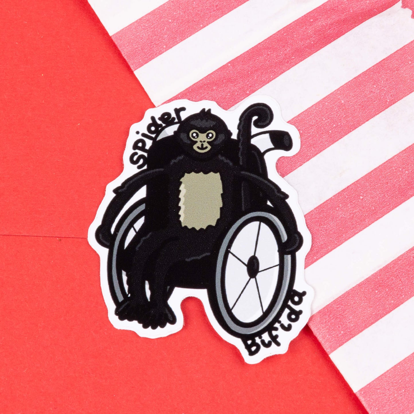 A cartoon sticker of a black spider monkey sitting in a wheelchair. The monkey has a light-colored patch on its chest and a smiling face. The text "Spider Bifida" is written on the sticker. The sticker is placed on a red background with a diagonal striped pink and white paper bag in the corner. Hand drawn design created created to raise awareness for spina bifida.