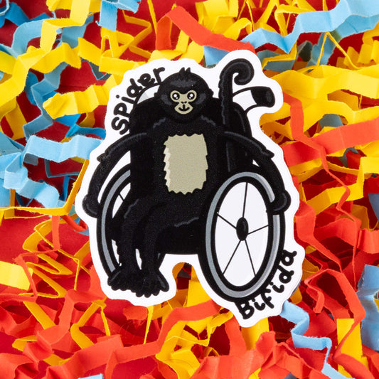 A cartoon sticker of a black spider monkey sitting in a wheelchair. The monkey has a light-colored patch on its chest and a smiling face. The text "Spider Bifida" is written on the sticker. The sticker is placed on a background of colorful shredded paper in yellow, red, and blue. Hand drawn design created created to raise awareness for spina bifida.