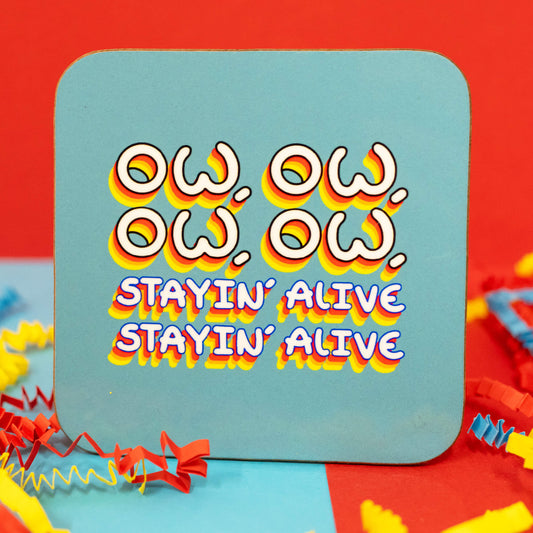 Close-up of the Ow, Ow, Ow, Ow, Staying Alive Coaster by Innabox. The square coaster has rounded corners and a light teal background. The design features the words "Ow, Ow, Ow, Ow" in bold white text with a vibrant red, yellow, and orange shadow effect, followed by "Stayin' Alive, Stayin' Alive" in blue and white retro-style font. Colourful red, blue, and yellow crepe paper surrounds the coaster.