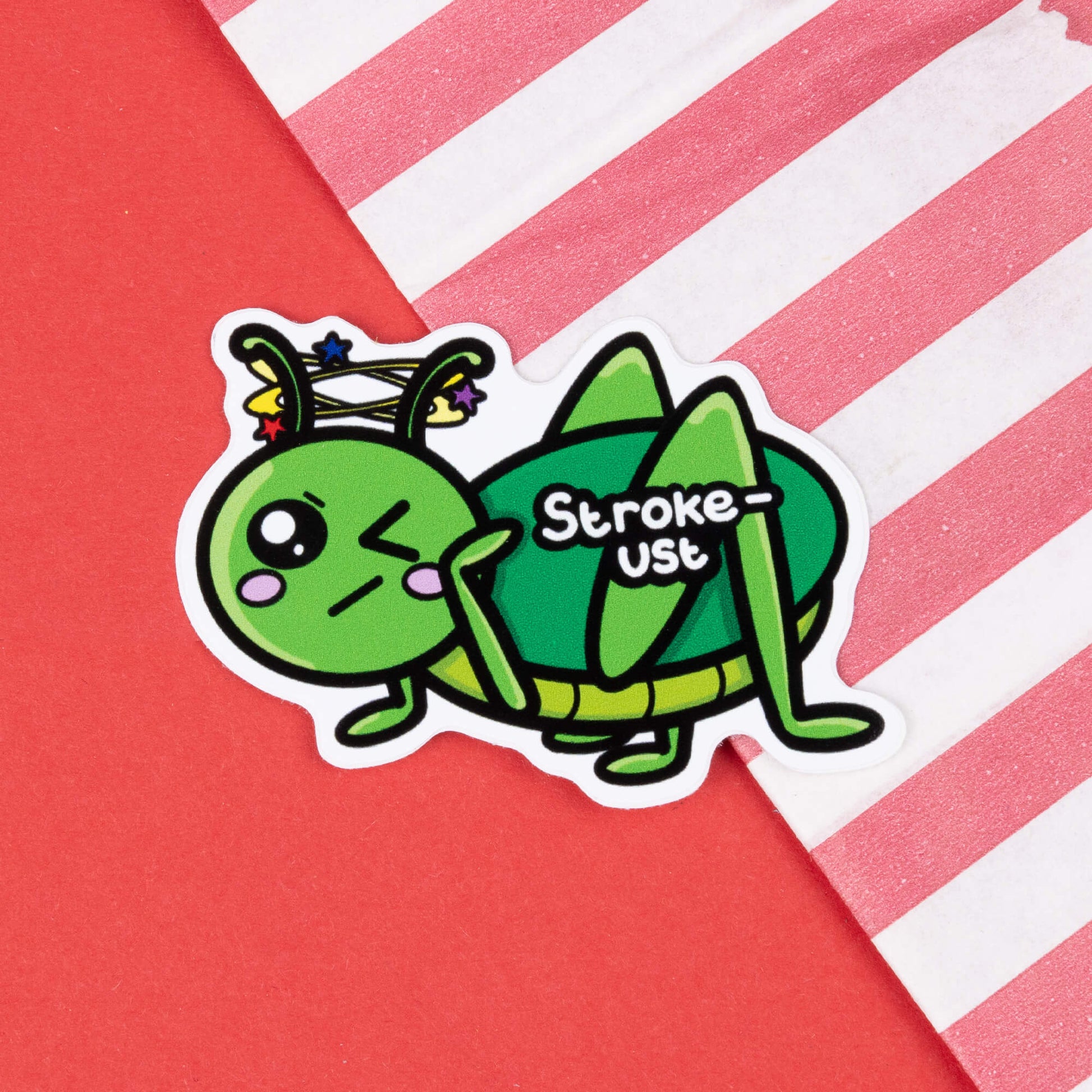 A cartoon sticker of a green grasshopper with a pained expression, blushing cheeks, and stars spinning around its head. The text "Stroke-ust" is written on the grasshopper's body. The sticker is placed on a red background with a diagonal striped pink and white paper bag in the corner. Hand drawn design created to raise awareness for strokes.