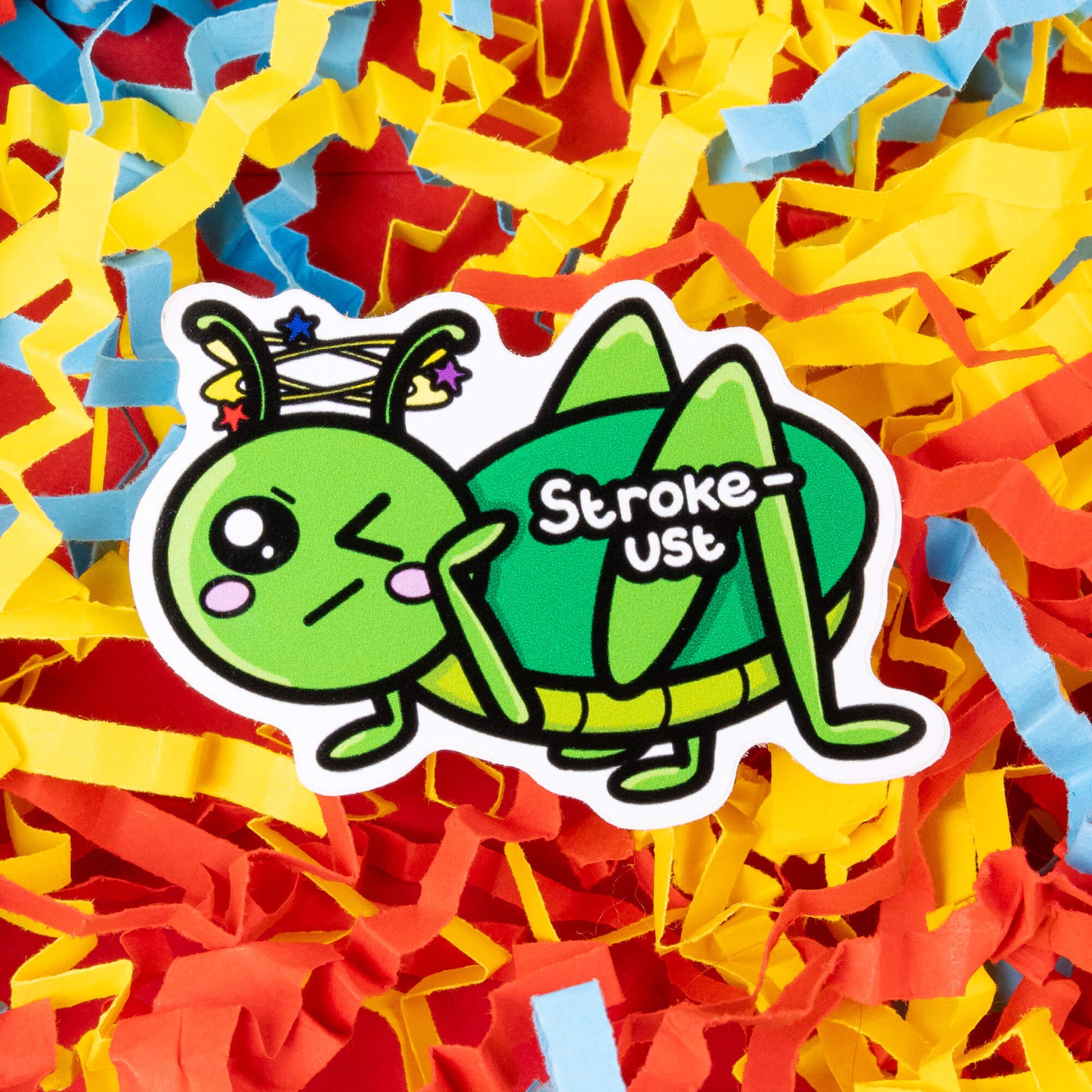 A cartoon sticker of a green grasshopper with a pained expression, blushing cheeks, and stars spinning around its head. The text "Stroke-ust" is written on the grasshopper's body. The sticker is placed on a background of colorful shredded paper in yellow, red, and blue. Hand drawn design created to raise awareness for strokes.