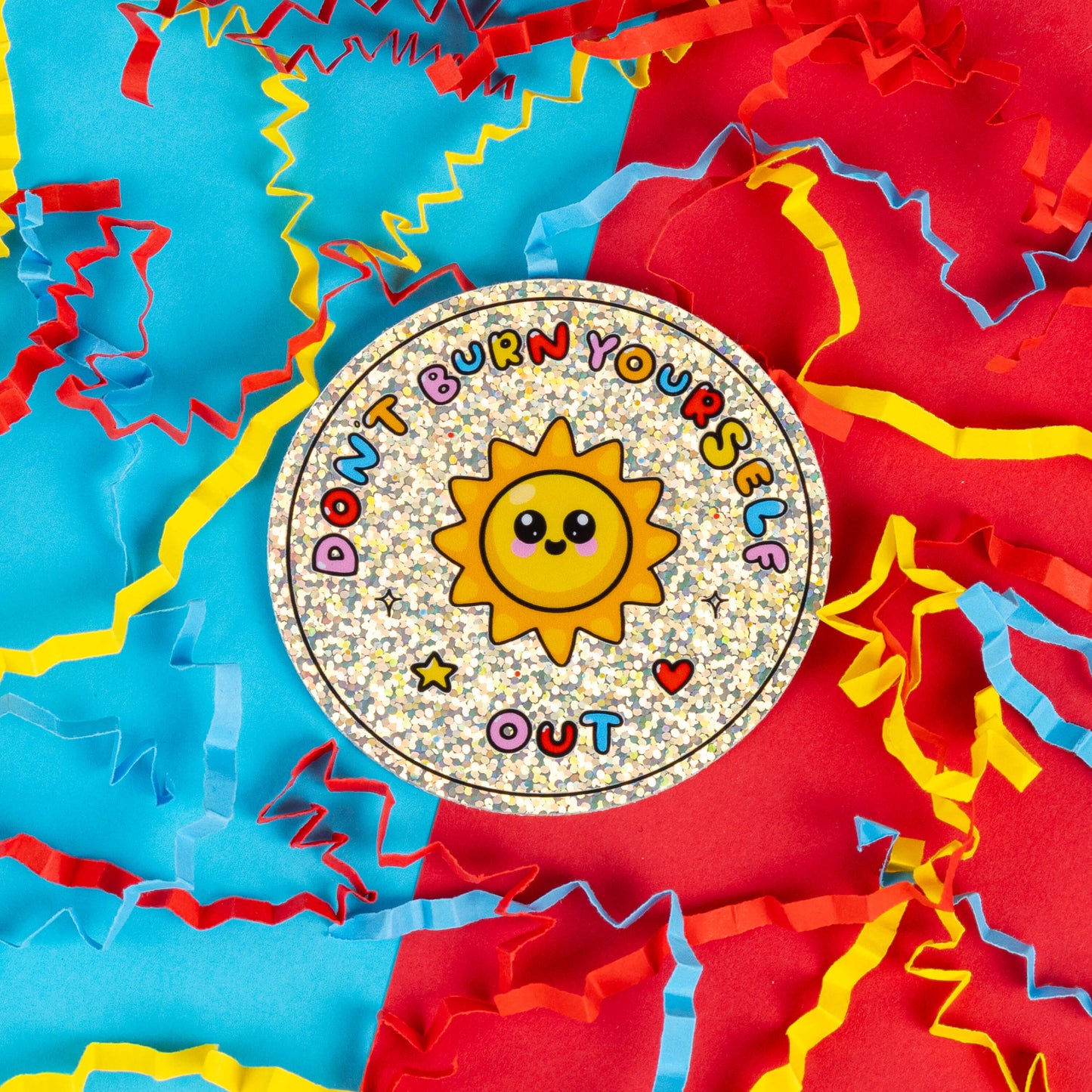 Innabox sticker featuring a smiling yellow sun with big, friendly eyes on a sparkling, holographic background. The text around the sun reads 'Don't Burn Yourself Out' in colourful, playful letters. Surrounded by vibrant red, blue, and yellow paper confetti, this sticker serves as a gentle reminder to take breaks and practise self-care.