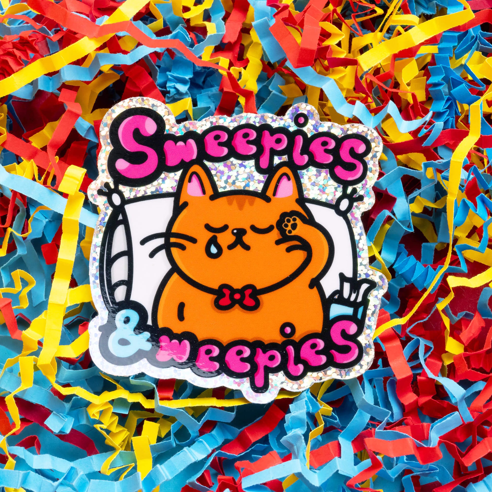 A close-up of an Innabox sticker showing an orange cat crying with the text "Sweepies & Weepies" in pink and black. The cat, wearing a red bowtie, holds a paw to its eye, symbolising the fatigue and emotional struggles faced by those with chronic illnesses. The background features a mix of light blue and red colours, creating a striking contrast. This disability-inspired design aims to offer comfort and validation to individuals managing chronic conditions.