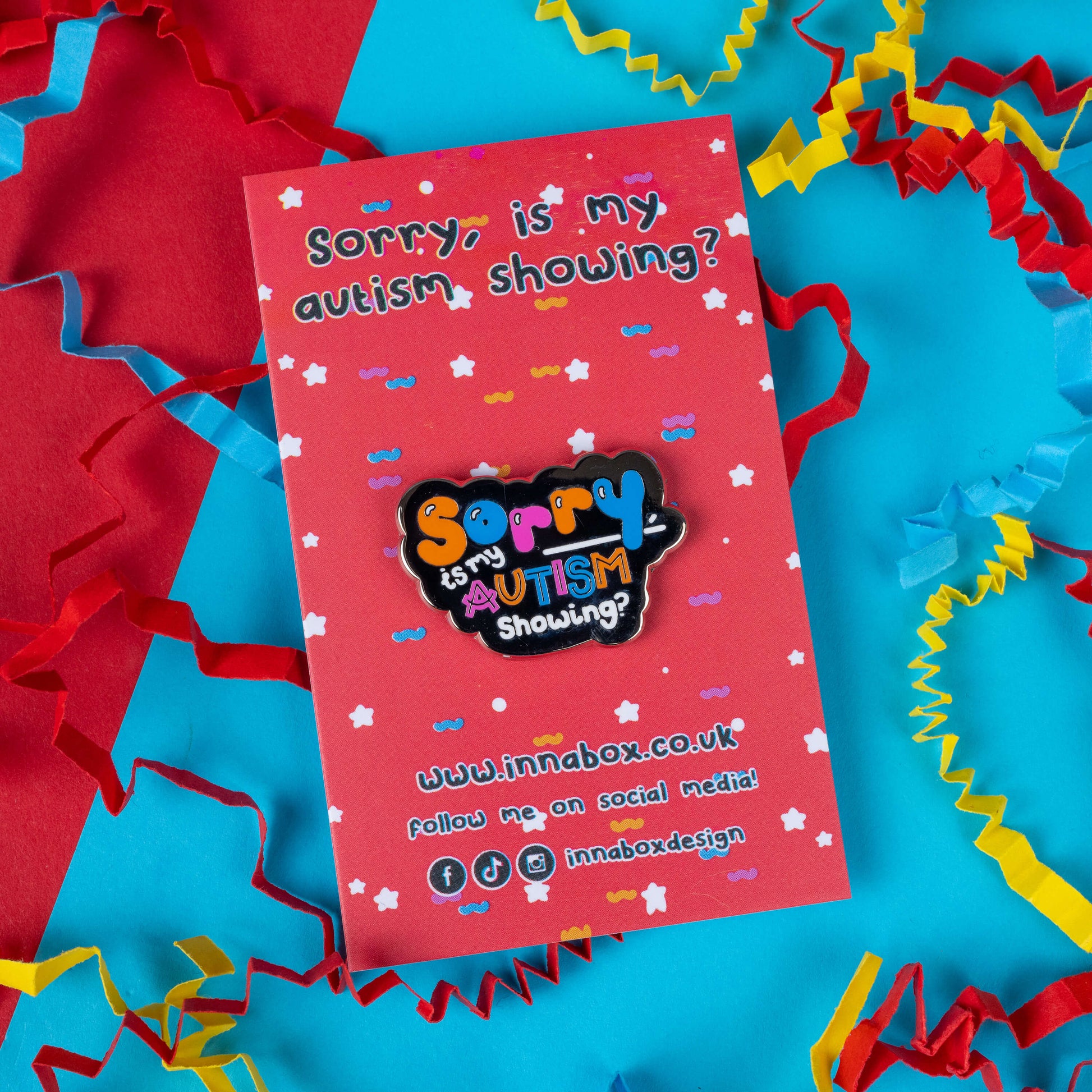 Innabox's 'Sorry, Is My Autism Showing?' enamel pin displayed on a red and blue background with colourful confetti paper. The pin features playful, multicoloured text and promotes autism awareness with a light-hearted message. Innabox creates uplifting disability-inspired products aimed at empowering those with chronic illnesses or hidden disabilities. Ideal for spreading awareness and adding personality to any outfit or accessory collection.