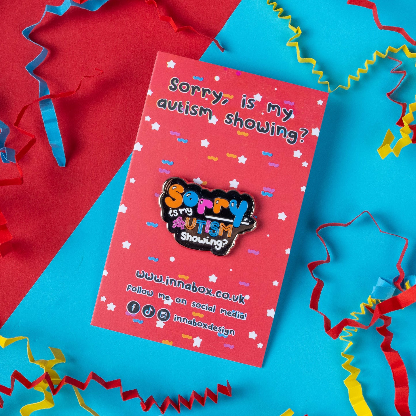 Innabox's 'Sorry, Is My Autism Showing?' enamel pin displayed against a colourful backdrop, with a red and blue background and paper confetti. The pin, with bright, cheerful lettering, raises autism awareness while adding a fun touch to any accessory. Created by Innabox, a brand dedicated to disability-inspired products to uplift individuals with chronic illnesses or hidden disabilities, this pin combines style and purpose for a meaningful accessory.