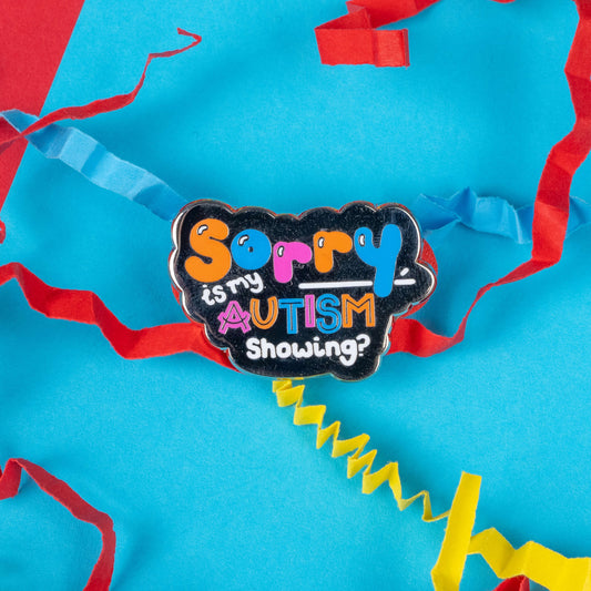 Innabox's 'Sorry, Is My Autism Showing?' enamel pin surrounded by colourful paper confetti on a vibrant blue and red background. The pin showcases multicoloured text in a playful font, sparking conversations around autism awareness. Innabox crafts disability-inspired products that uplift those with chronic illnesses and hidden disabilities, making this pin a fun yet impactful way to promote understanding and acceptance.