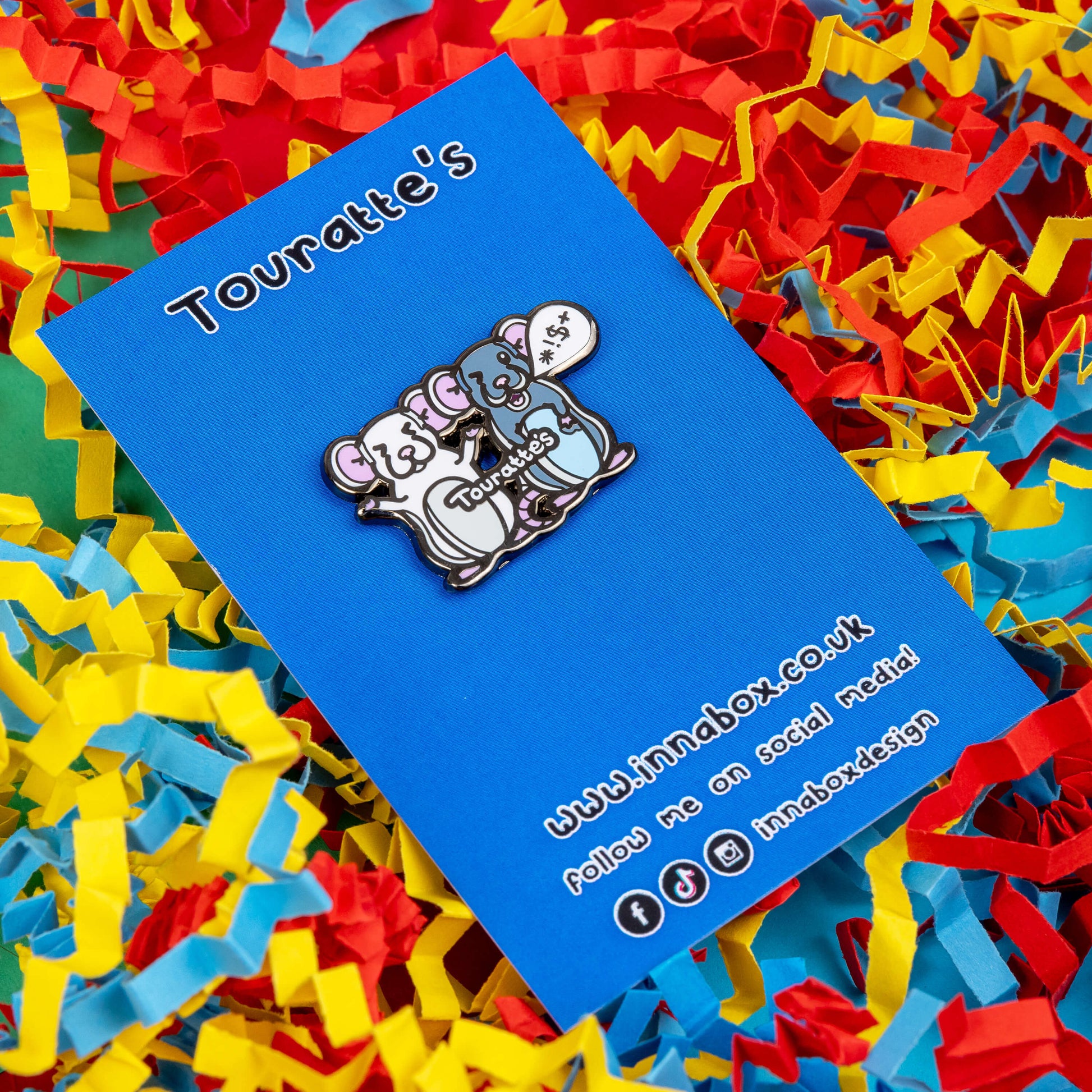 The Touratte's Rat Enamel Pin - Tourette's Syndrome on blue backing card laying on a red, blue and yellow paper background. The enamel pin is of two rats with pink ears and feet, the white rat on the left is raising its arms with its eyes closed and the grey rat on the right has a speech bubble with symbols in, across both rats reads 'touratte's'. The hand drawn design is raising awareness for tics and tourettes.