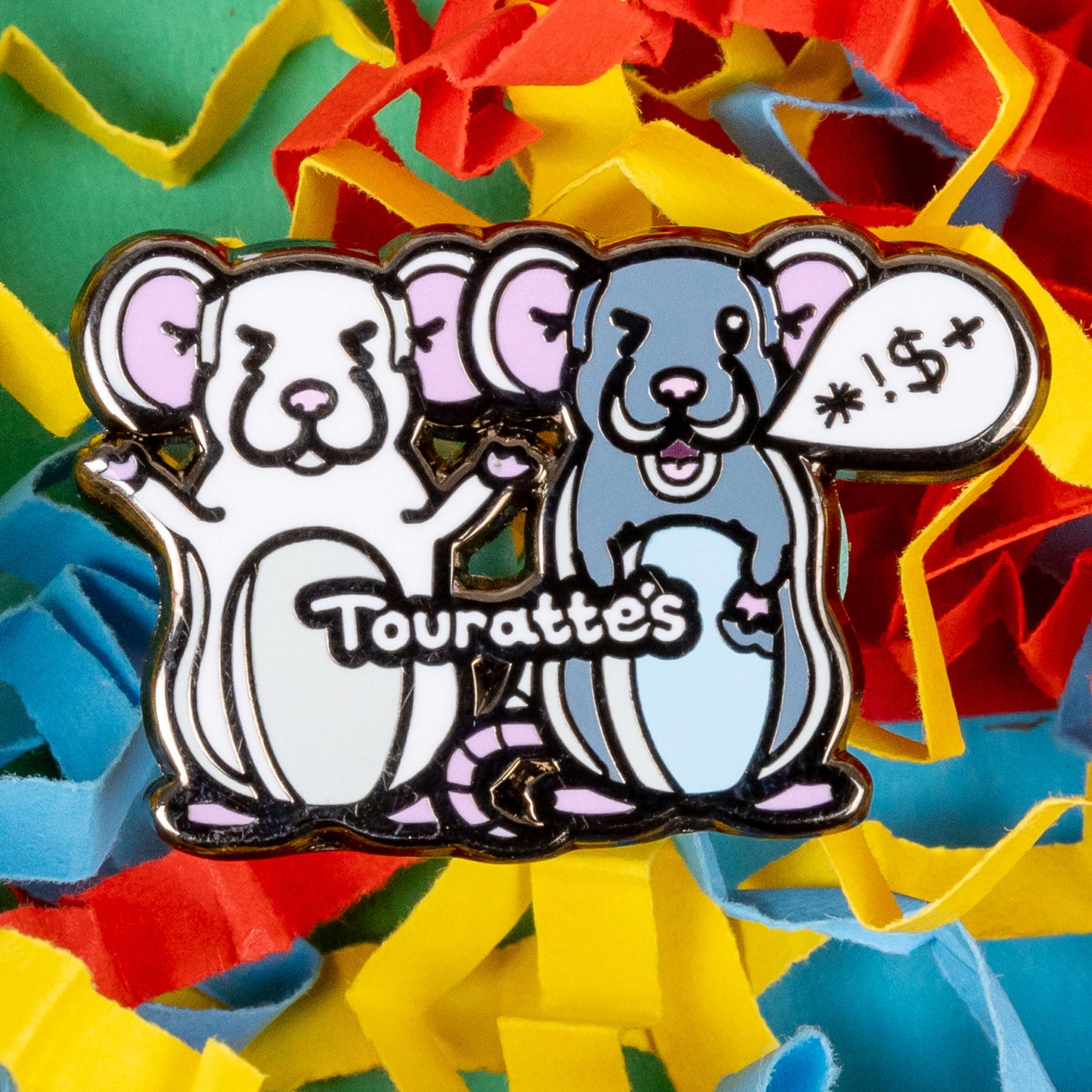 The Touratte's Rat Enamel Pin - Tourette's Syndrome on a red, blue and yellow paper background. The enamel pin is of two rats with pink ears and feet, the white rat on the left is raising its arms with its eyes closed and the grey rat on the right has a speech bubble with symbols in, across both rats reads 'touratte's'. The hand drawn design is raising awareness for tics and tourettes.