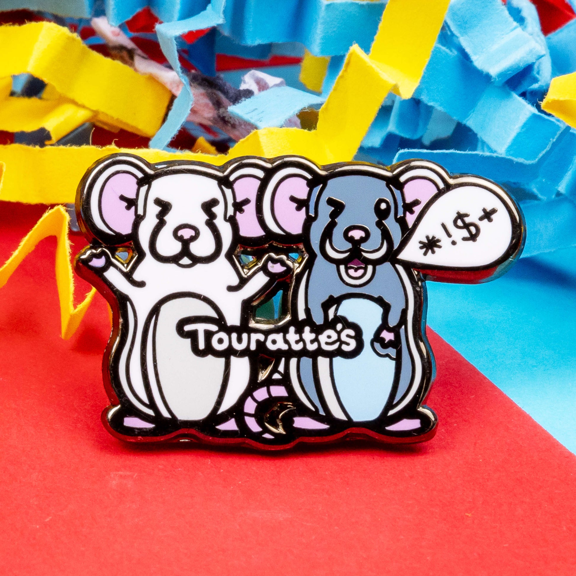 The Touratte's Rat Enamel Pin - Tourette's Syndrome on a red, blue and yellow paper background. The enamel pin is of two rats with pink ears and feet, the white rat on the left is raising its arms with its eyes closed and the grey rat on the right has a speech bubble with symbols in, across both rats reads 'touratte's'. The hand drawn design is raising awareness for tics and tourettes.