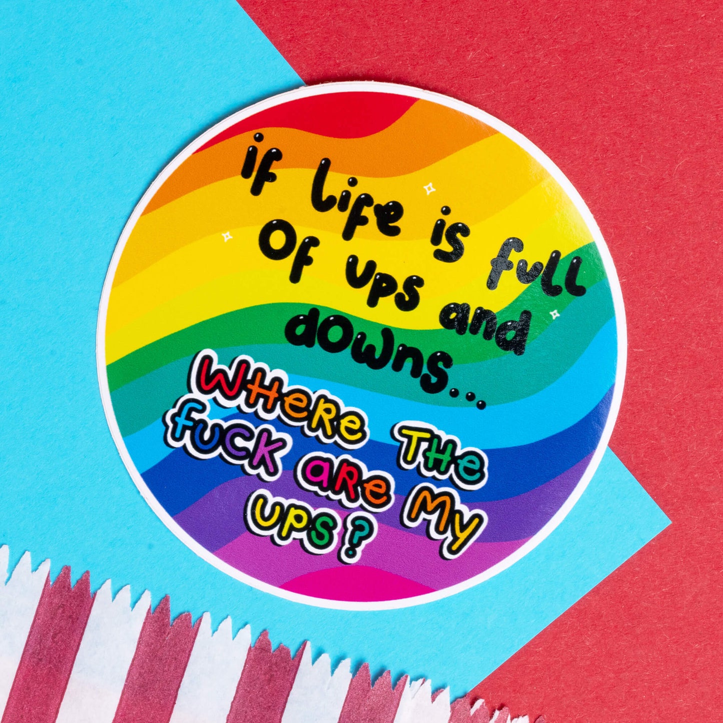 Colourful circular sticker by Innabox with rainbow stripes, featuring bold text: 'If life is full of ups and downs... Where the f**k are my ups?' This uplifting design reflects the frustration of those with chronic illnesses or hidden disabilities, blending humour and resilience. Set against a bright background of red and blue papers, it adds vibrancy to disability awareness.