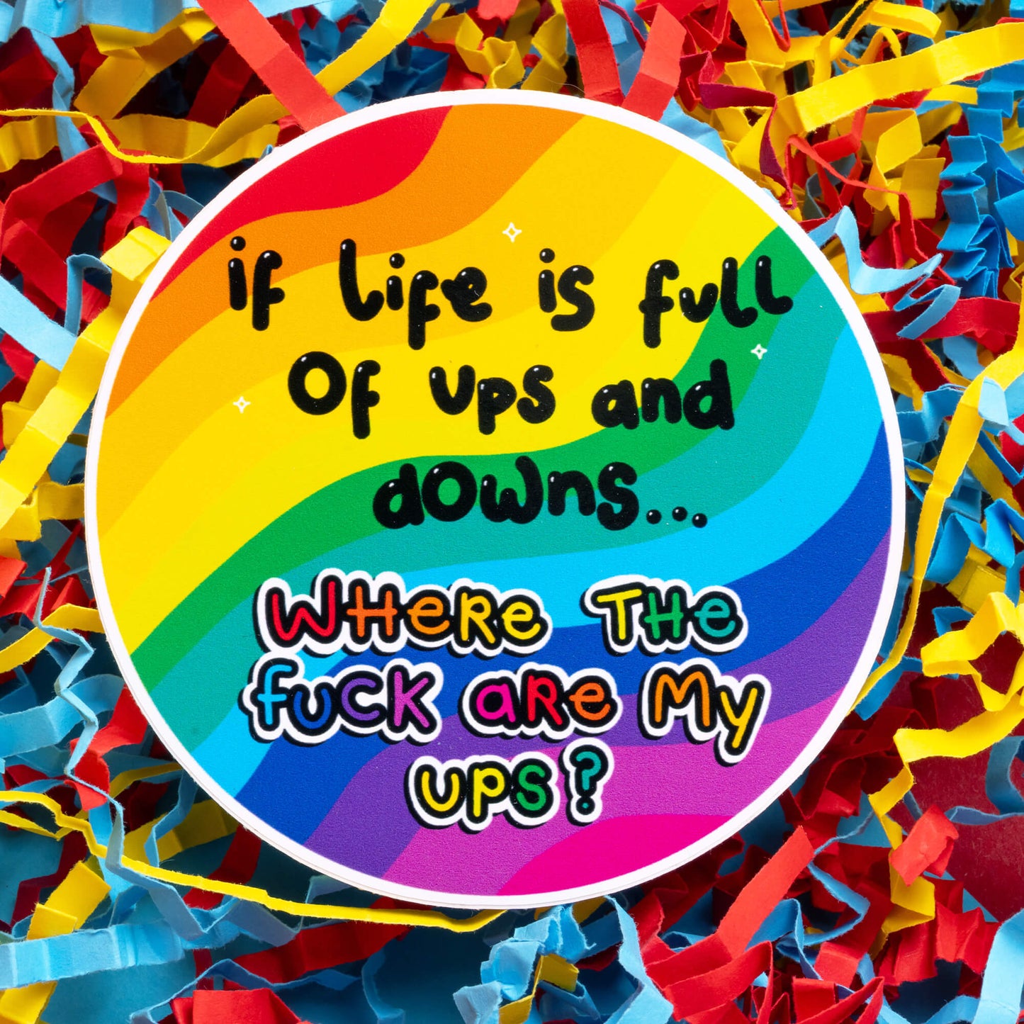 Innabox sticker with rainbow colours and the words: 'If life is full of ups and downs... Where the f**k are my ups?' Designed for those with chronic illnesses or hidden disabilities, it offers humour and support. Displayed on a background of colourful shredded paper, symbolising the chaotic yet vibrant journey of living with disability.