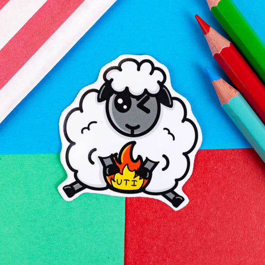 The Ewe TI - UTI Sticker on a red, blue and green background with colouring pencils and red stripe candy bag. The ewe sheep shaped sticker has one eye scrunched shut with its front hooves over its lower belly with an orange and yellow fire with black text reading 'UTI'. The hand drawn design is raising awareness for urinary tract infections.